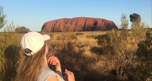 Au pair, mit dem Rücken zur Kamera, schaut auf den Uluru in der Ferne